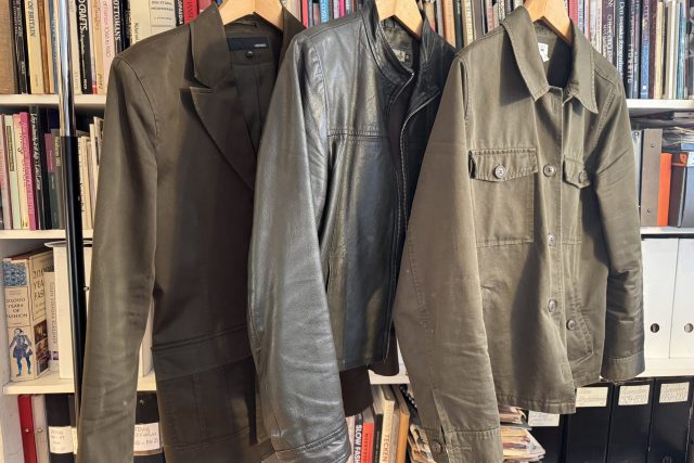 Tre olivgröna kavajer - en blazer, en skinnjacka och en bruksjacka - hänger på trägalgar framför en bokhylla fylld med böcker och arkivlådor.