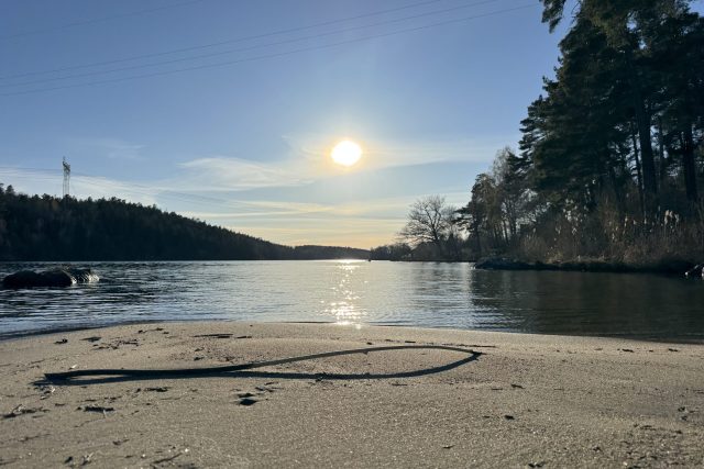 En lugn flod i solnedgången med solen lågt på himlen som ger reflexer i vattnet. Sandstrand i förgrunden, omgiven av skog på båda sidor.