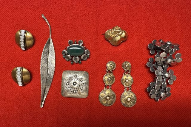 En samling vintagesmycken, bl.a. örhängen, broscher och pins i guld- och silvertoner med olika mönster, på en ljusröd tygbakgrund.