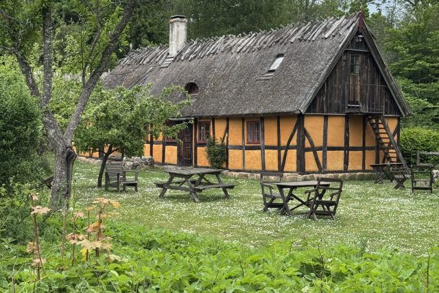 En pittoresk gul korsvirkesstuga med halmtak ligger i en grönskande trädgård, omgiven av träd och flera picknickbord i trä på gräset.