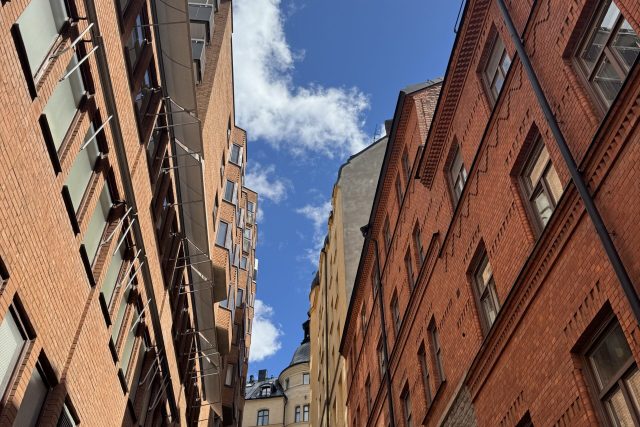 En smal trappa reser sig mellan höga röda tegelbyggnader under en ljusblå himmel med spridda vita moln. Lampor lyser i trappan och några personer syns högst upp.